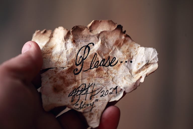 A hand holds an aged, crumpled paper with a handwritten note saying 'Please' in cursive.