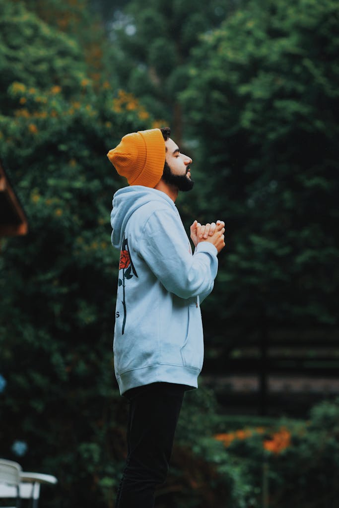 A man in a beanie and hoodie deeply praying outside in a lush green forest.