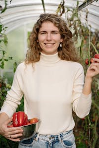 A smiling woman holds freshly harvested bell peppers inside a lush greenhouse.