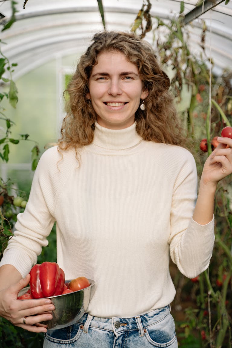 A smiling woman holds freshly harvested bell peppers inside a lush greenhouse.