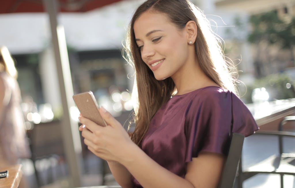 A smiling young woman in a purple top texting on her smartphone outdoors.