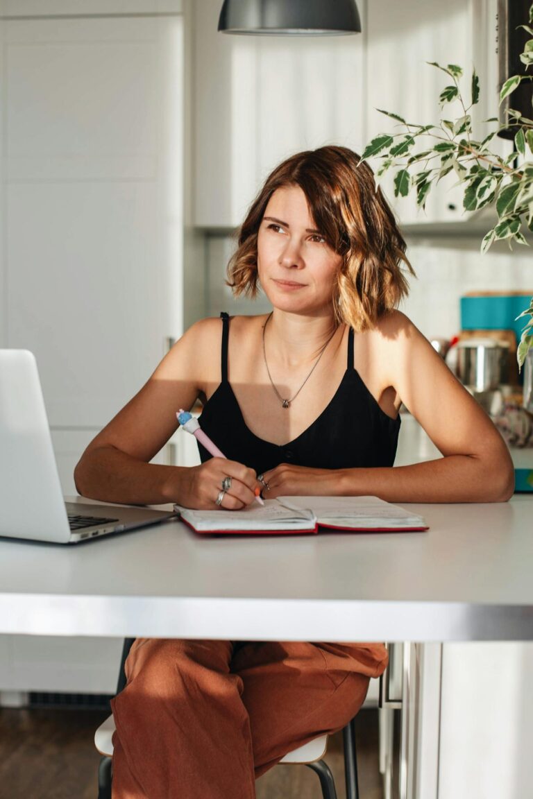 A woman in a black tank top writing with a laptop at a home desk during the day.