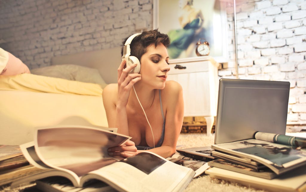 A woman with headphones enjoys music while surrounded by magazines and a laptop in a cozy bedroom setting.
