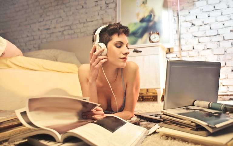 A woman with headphones enjoys music while surrounded by magazines and a laptop in a cozy bedroom setting.