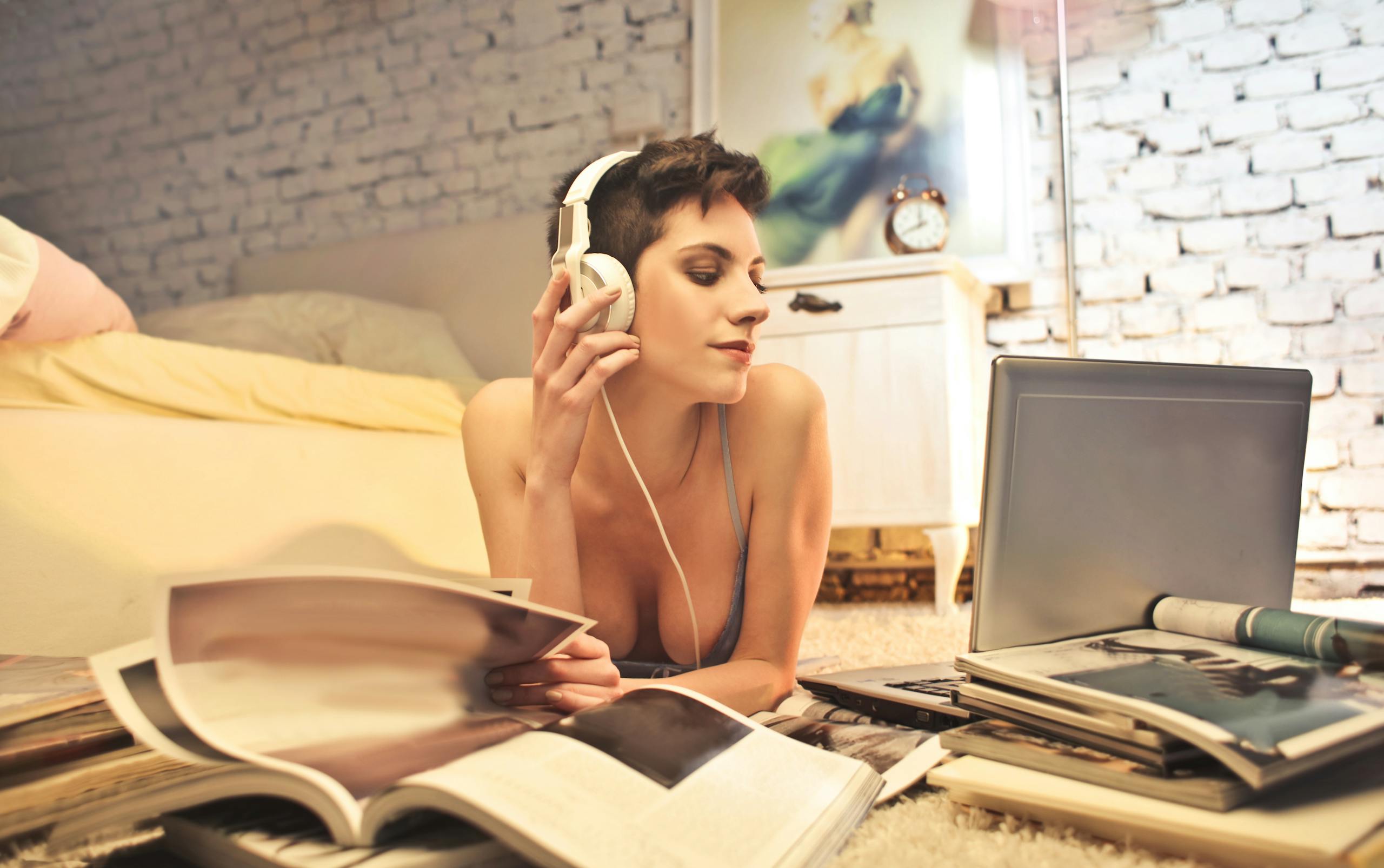 A woman with headphones enjoys music while surrounded by magazines and a laptop in a cozy bedroom setting.
