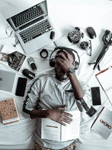 A young man lying on a bed surrounded by modern gadgets, books, and tech devices, illustrating digital lifestyle.
