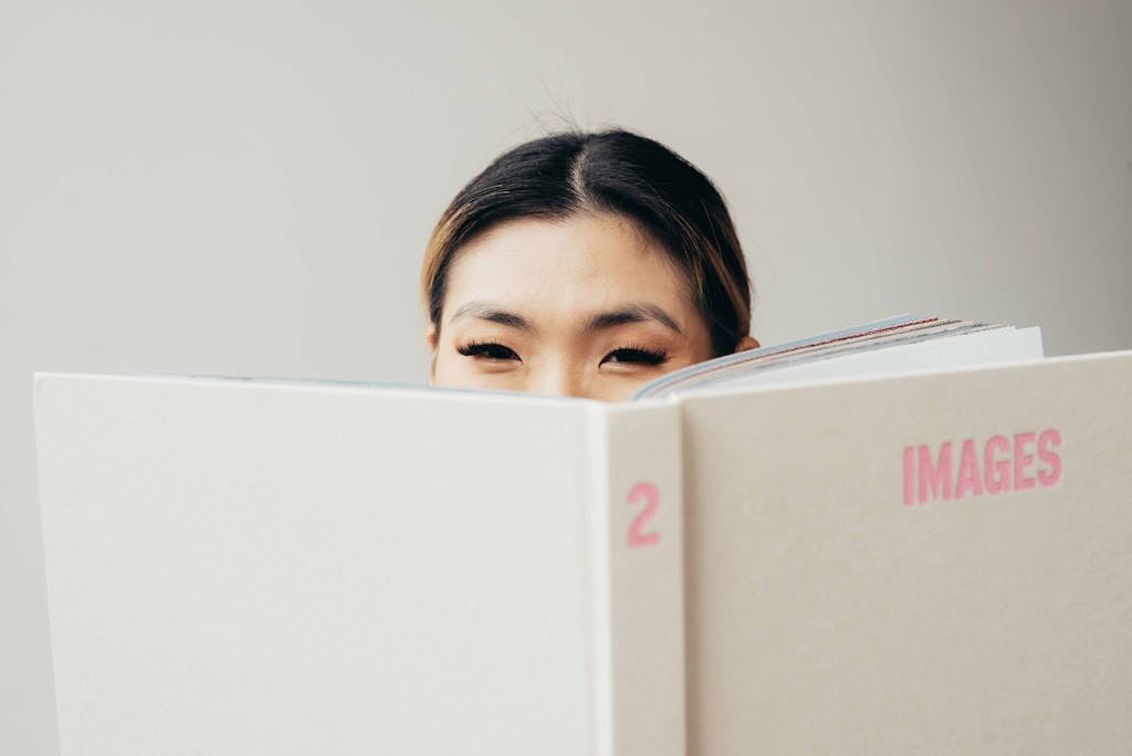 A young woman peeks over a large book, creating a playful and engaging image for stock use.