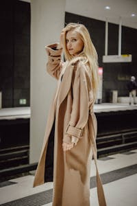 Fashionable woman models a beige coat while waiting at a railway station platform.