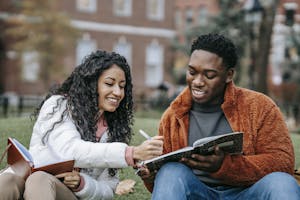 Happy college students studying together on a campus lawn, sharing ideas and writing notes.