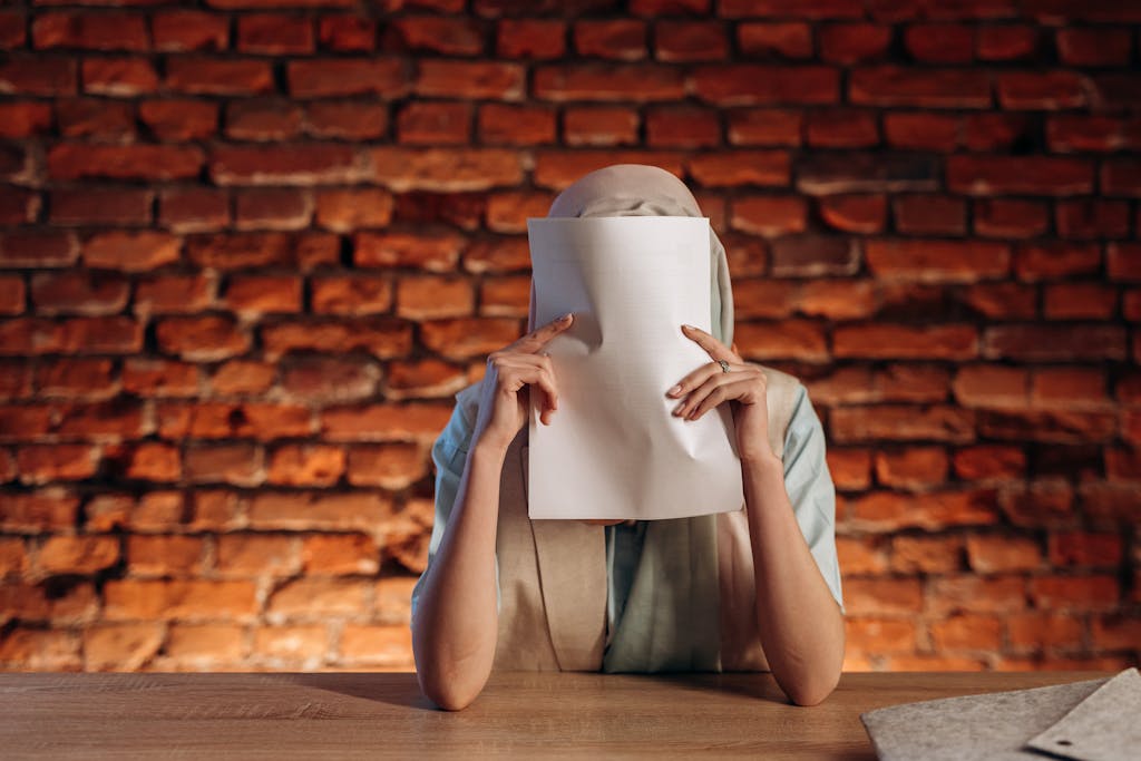 Individual at desk holds paper, obscuring face against a textured brick wall.