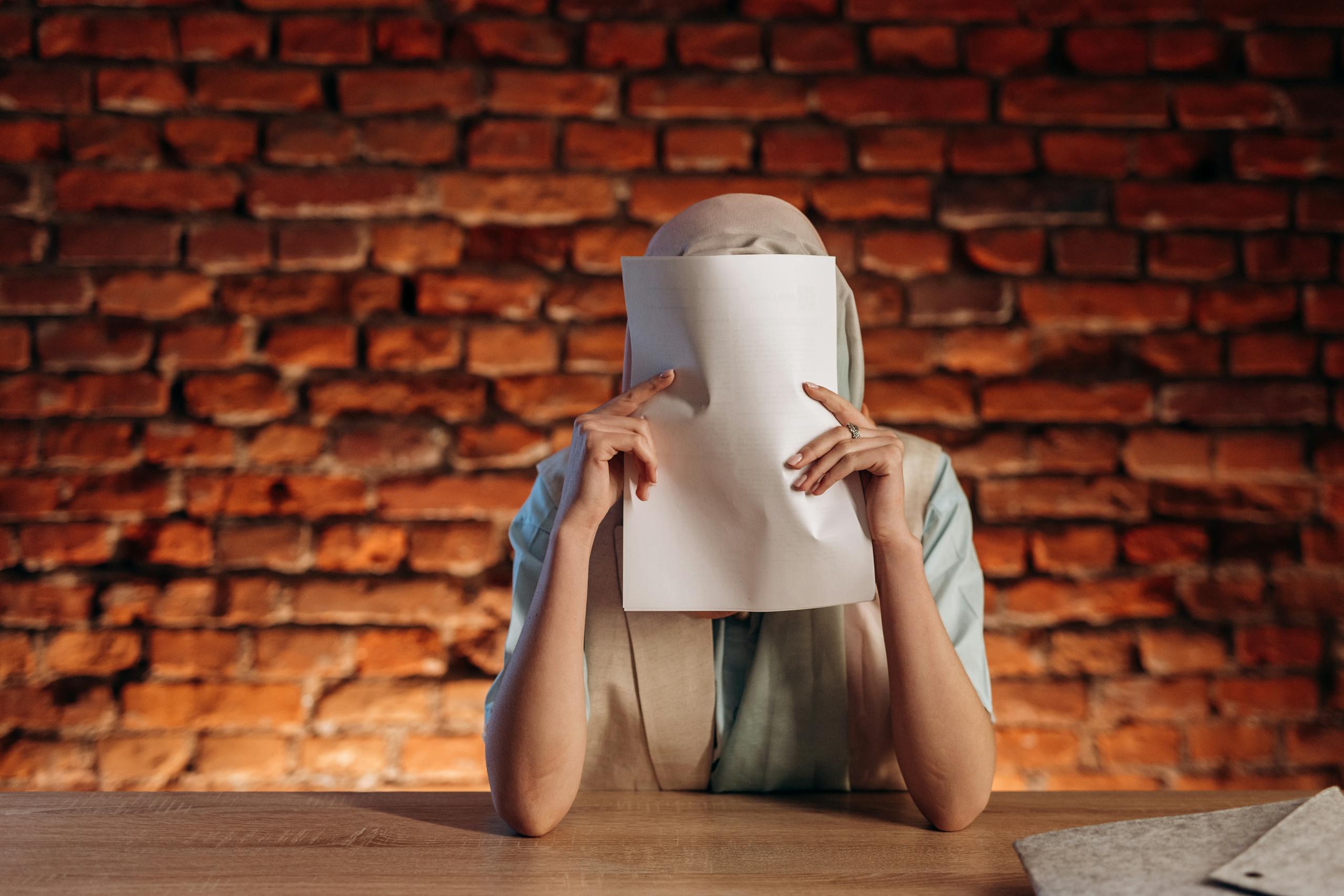 Individual at desk holds paper, obscuring face against a textured brick wall.