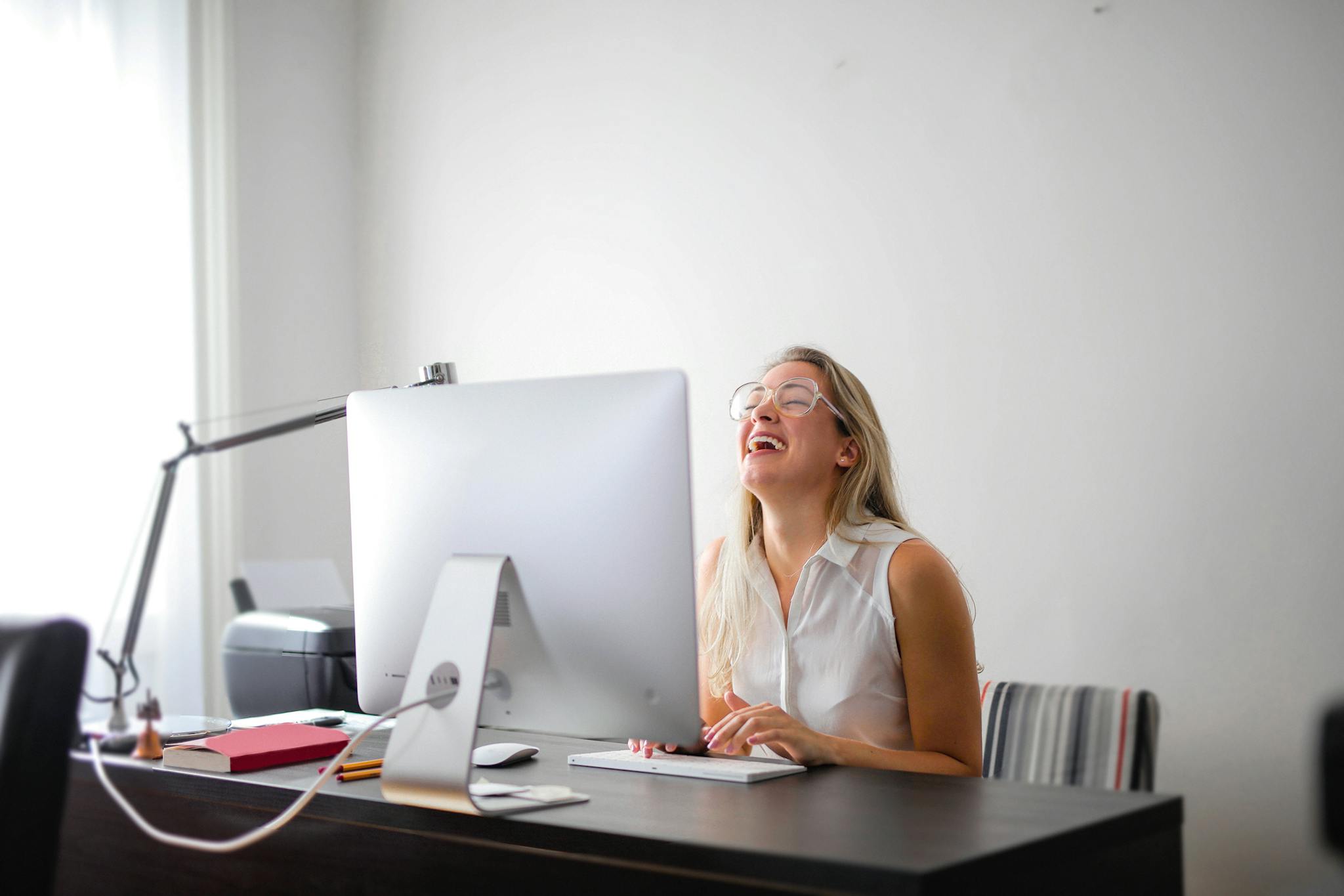 Office scene with a woman laughing while working at a modern desk, showcasing joy and productivity.