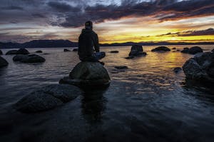 Silhouette of a man seated on rocks, gazing at a serene Lake Tahoe sunset.