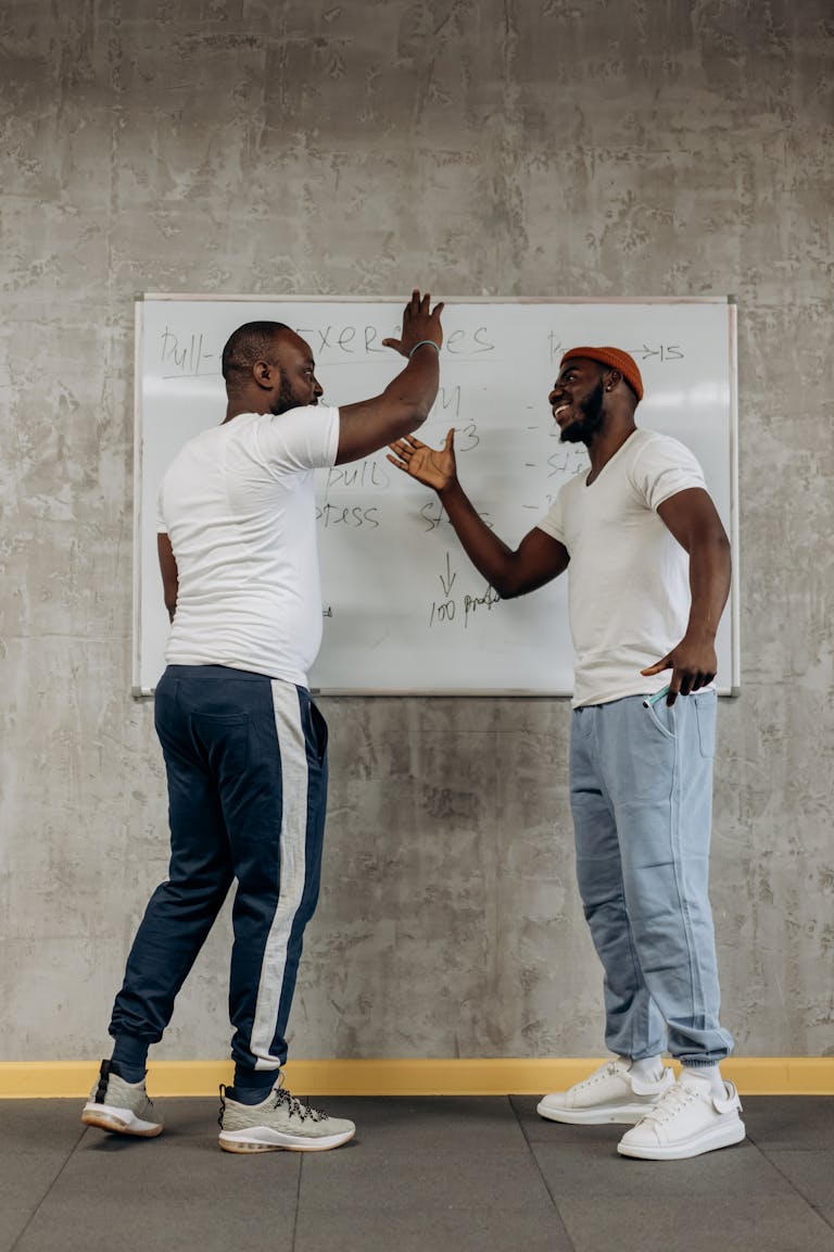 Two men celebrate with a high five in a gym, planning a workout session on a whiteboard.