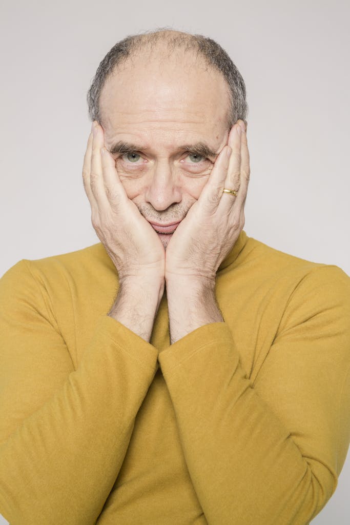 Elderly man in yellow sweater posing with hands on face, studio portrait.
