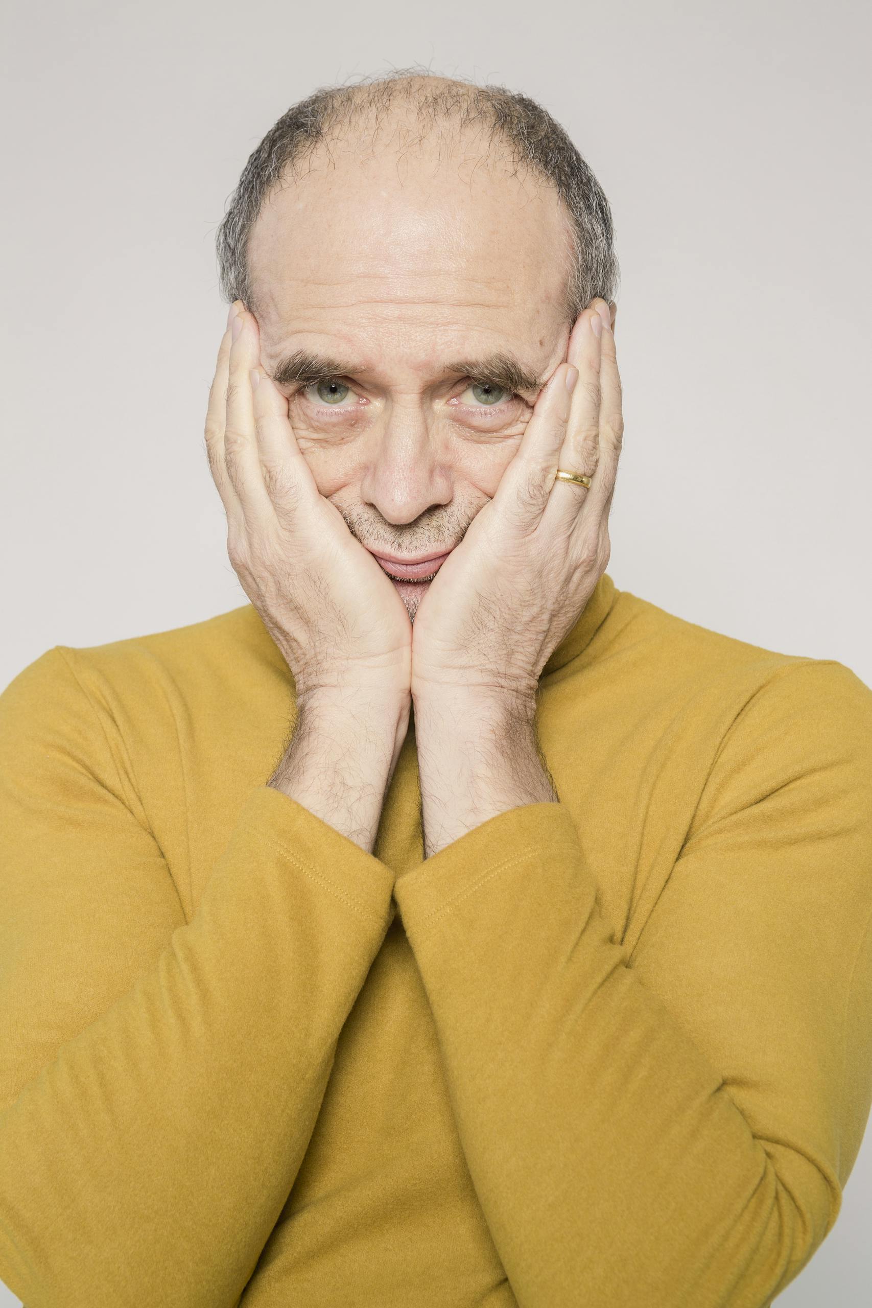 Elderly man in yellow sweater posing with hands on face, studio portrait.
