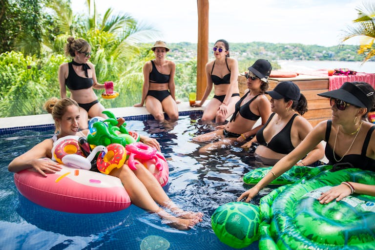 Seven women in bikinis relaxing and having fun at an outdoor pool party.