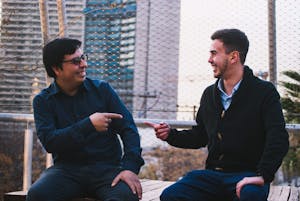 Two men sitting outdoors, smiling and pointing at each other in a friendly gesture.