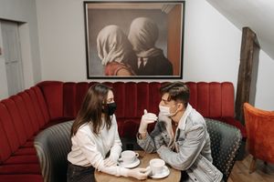 A couple enjoys coffee while wearing masks in a cozy caf&eacute;, highlighting the pandemic's impact.