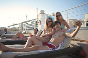 Group of young happy ladies and guy in swimsuits and sunglasses sitting on deck chair and taking selfie while chilling together during summer vacation